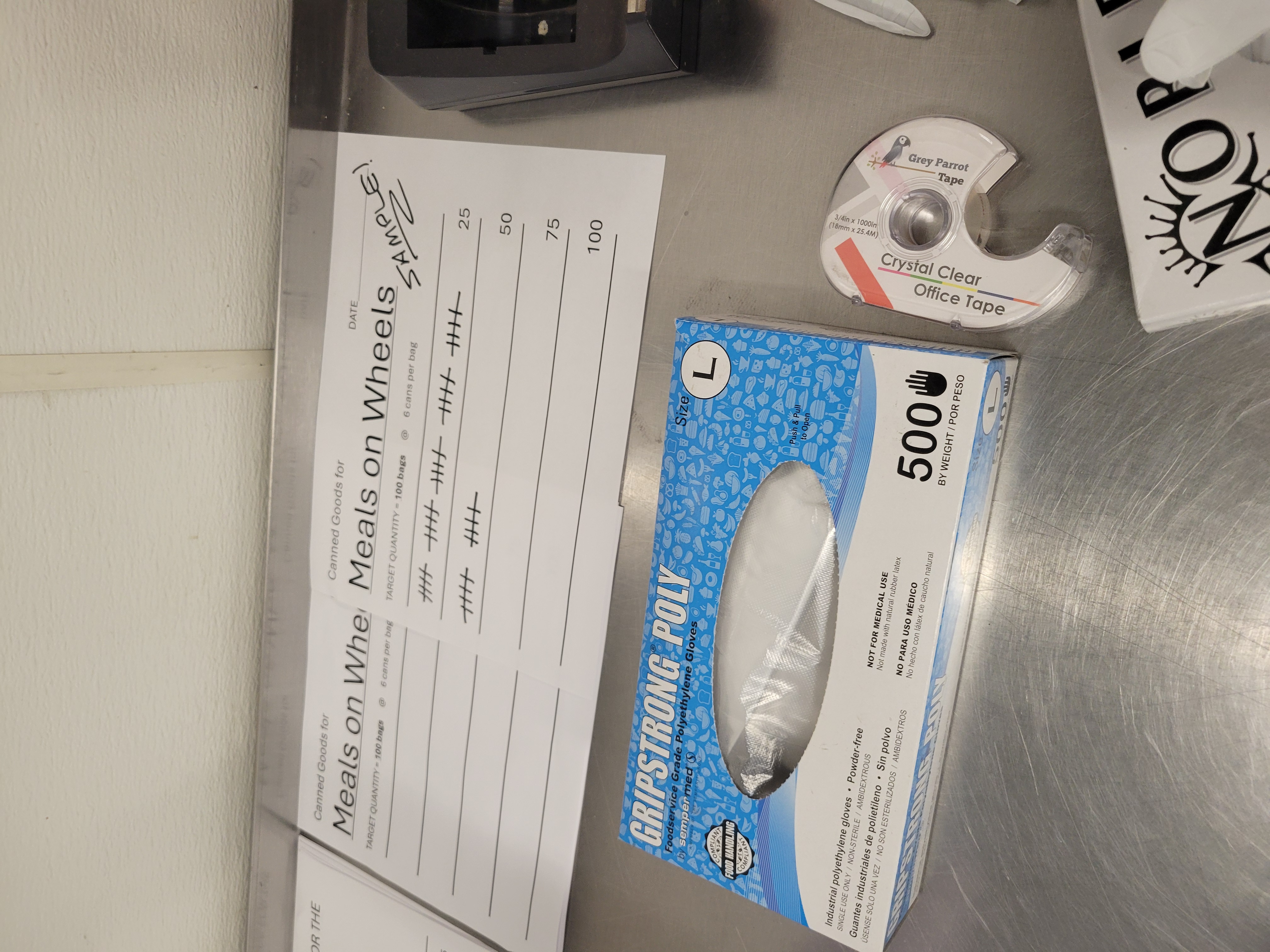 Piece of paper listing the tally marks of 100 bags of 6 canned goods each for Meals on Wheels on a table with a box of gloves and tape in front of it.